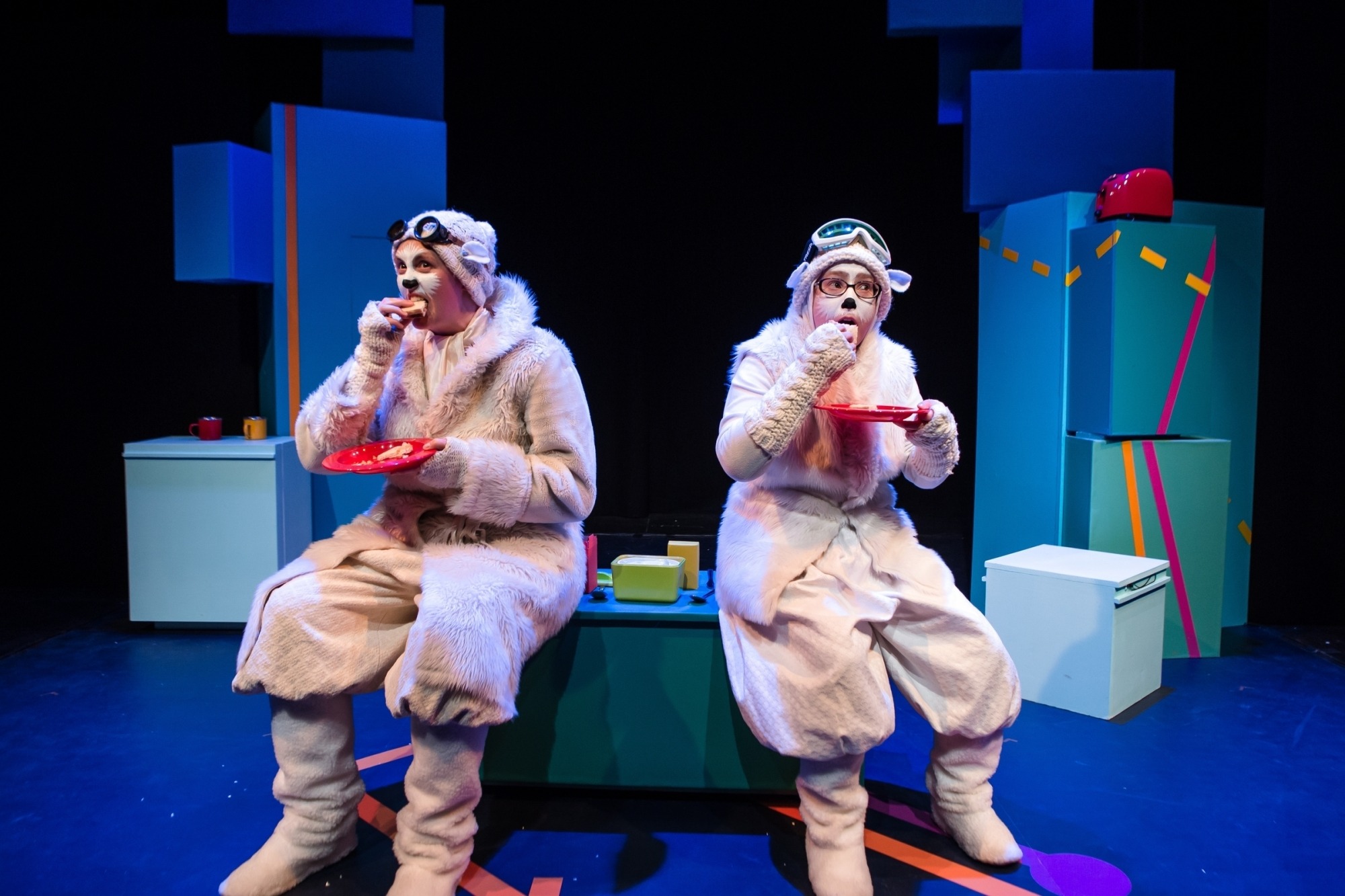 Image of two characters from The Polar Bear Goes Up dressed in polar bear costumes eating sandwiches off of red plates while sitting on a blue set
