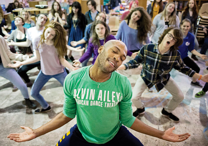Image of a dancer in a green Alvin Ailey T-shirt facing the camera leading a dance master class for UNL students behind him