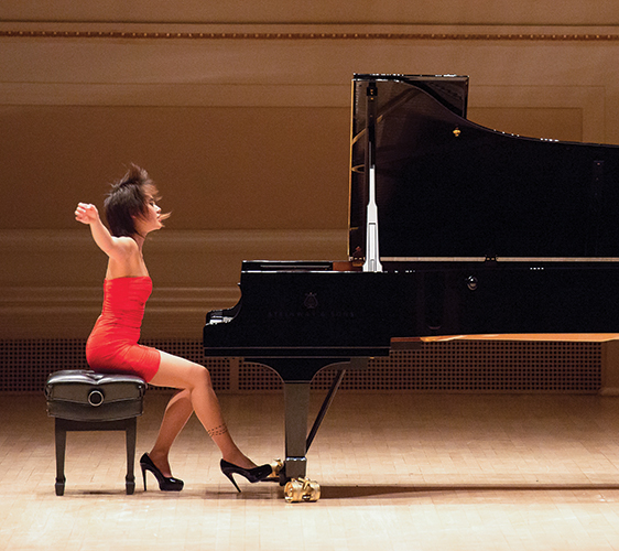 Image of Yuja Wang seated at a piano with arms raised and hair flying forward