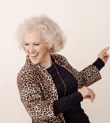 a woman wth short curly blonde hair and a cheetah print jacket smiles and dances