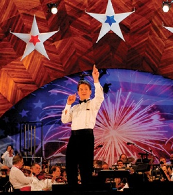 A man in a white shirt and black pants facing the audience with an orchestra behind him. The stage has red, white and blue stars