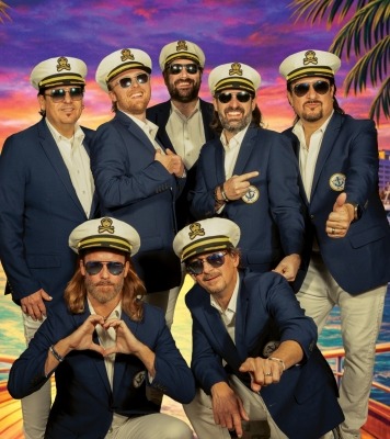 a seven-piece yacht rock band poses in captains outfits against a vibrant ocean sunset