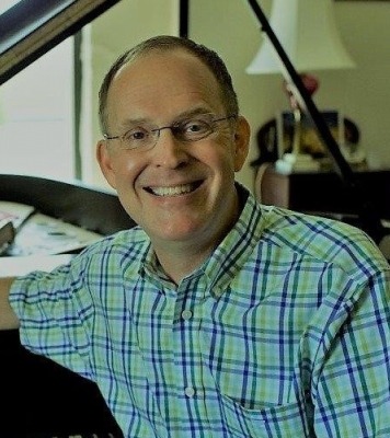 a man with glasses casually sits at a piano in a blue and green plaid button up shirt