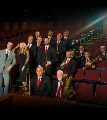 Orchestra members in theater seats holding their instruments and smiling at the camera
