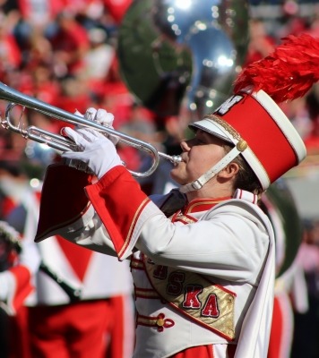 A student stands in the forefront in a bright red and white cornhusker marching band uniform playing a trumpet towards an off-screen crowd. 