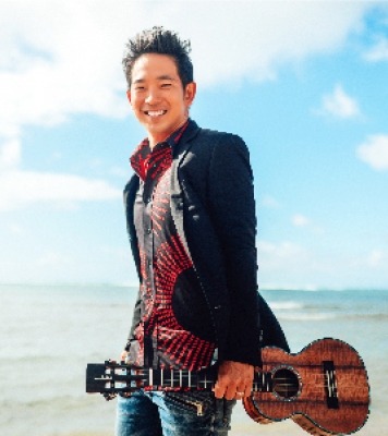 Man with dark short hair standing on a beach in a suit jacket and red and black shirt, holding a ukulele. The background shows the sky with clouds and the ocean. 