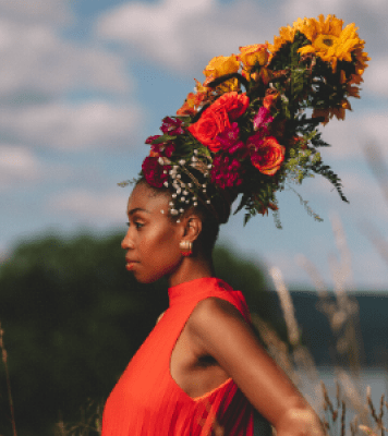 Picture of women with red dress on looking away from the photo. She is wearing a tall hat of multiple different flowers. She is standing in a field.