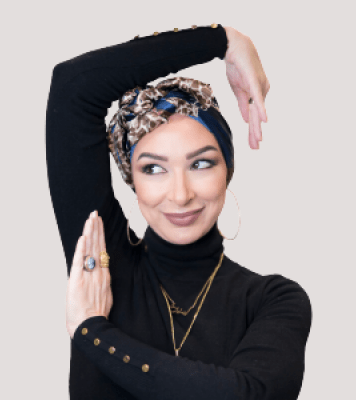 Woman faces the camera smiling. She has her arms raised and posed around her head. She wears gold jewelry and a black turtleneck sweater. 