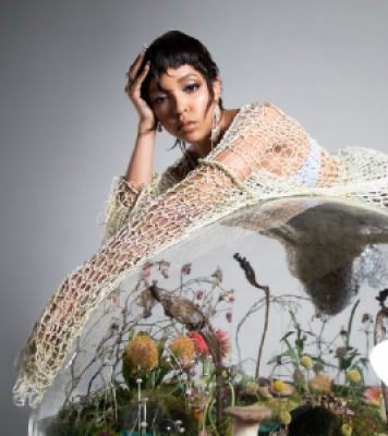 Image of a girl with short, dark hair laying across a glass ball terrarium. She is wearing a cream cover-up with a white top under it. She is looking towards he camera.