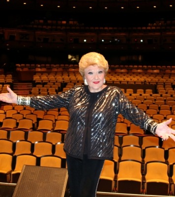 Woman in sparkly black jacket standing in front of theater seats with arms extended as if to welcome you to the theater