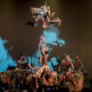 One man is flipping in the air after being thrown by another man. Another man watches and three drummers play in the background.