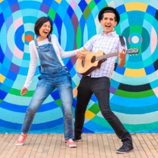 A woman in overalls and a man with a hat playing a guitar standing by each other in front of a colorful wall