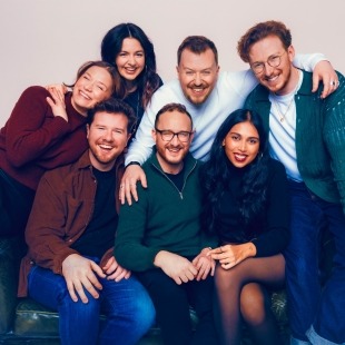 a group of seven smiling humans pose closely together on a small green loveseat