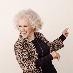 a woman wth short curly blonde hair and a cheetah print jacket smiles and dances