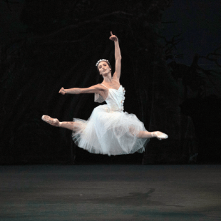 A ballerina in all white jumps into the splits in air contrasted against a black background 