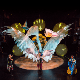 Large white crane puppet on stage. Two people holding the puppet up with strings. One person holding a smaller crane puppet at the front of the stage. Other puppets are showcased on the stage and lights are on the back wall of stage.