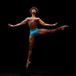 Lone man wearing blue shorts floats in the air against a black background, arms outstretched and one leg outstretched to one side
