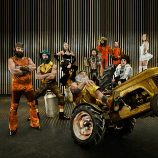 A group of nine people in colorful costumes face the camera. In the forefront and older man does a wheelie on a tractor, behind him, people stand in a variety of poses 