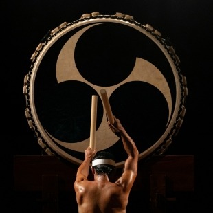 a shirtless man drums a very large drum with a graphically printed head