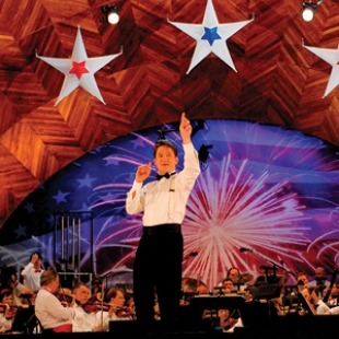 A man in a white shirt and black pants facing the audience with an orchestra behind him. The stage has red, white and blue stars