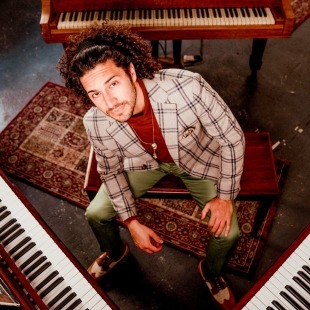 a man with curly hair and a funky suit sits between three pianos