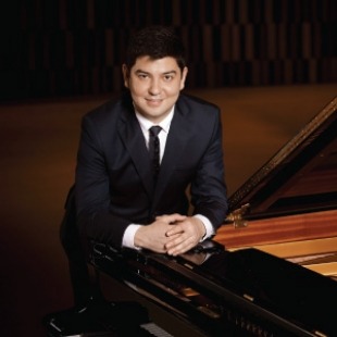 Man with dark hair in a suit and tie leaning forward onto a piano with his hands together.