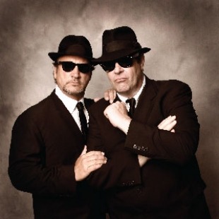 Two men in black suits, sunglasses, and hats against a dark background 