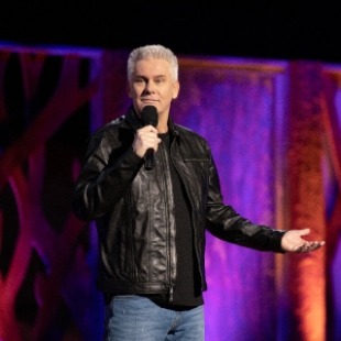 Man with white hair wearing a black shirt and black leather jacket along with blue jeans. He is holding a microphone to his mouth with his hand to the right out to the side. Standing behind a purple and orange backdrop. 