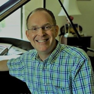a man with glasses casually sits at a piano in a blue and green plaid button up shirt