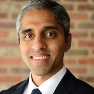 a smiling man in a classy suit gazes into the camera against a brick wall background