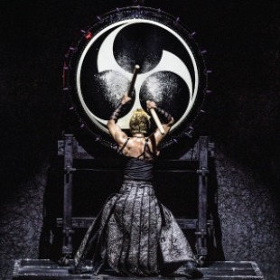 Person facing away from the camera in a black leather skirt and black sleeveless top. Arms are above their head holding two drum mallets. Banging the mallets on a large black drum with a white design on it.