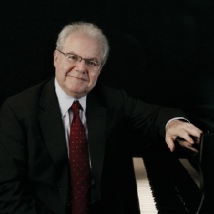 Male with white hair in suit with a red tie leaning on a piano with a black background. 
