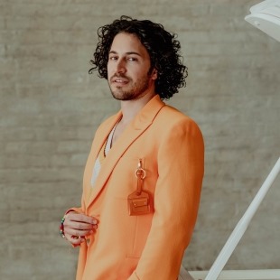 a man with curly brown hair and an orange fashion suit poses for photo