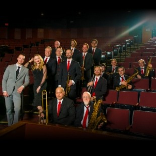 Men in black suits and red ties and a lady with a black dress and a man in a grey suit sitting and standing in red theatre seats. 
