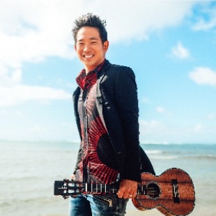 Man with dark short hair standing on a beach in a suit jacket and red and black shirt, holding a ukulele. The background shows the sky with clouds and the ocean. 