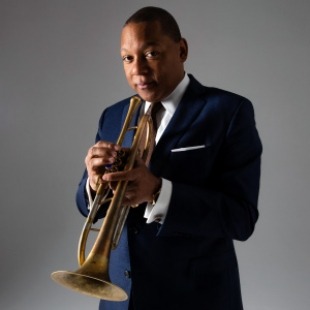 Man in a navy suit holding a trumpet close to his mouth standing in front of a grey background. 