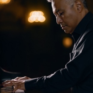 Man here a black long sleeve button down playing the piano. Lights are shining on him in the background.