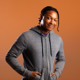 a man poses in a grey hoodie with hands in pockets and a cheeky smile