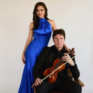 Man with short brown hair sitting in a chair wearing a black suit and black shirt holding a violin in his lap and holding a violin bow. Women with dark long hair standing behind him in a behind him in a long bright blue sleeveless dress with her hand on his shoulder.