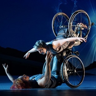 Alice Sheppard, a multiracial Black woman with coffee-colored skin and short curly hair, and Laurel Lawson, a white person with cropped hair, are perched in their wheelchairs on the edge of a huge ramp, backs exposed and sculpted in the light. Above them, bursts of light shine like spirits in a starry blue sky. Photo Jay Newman/BRITT