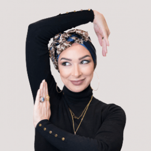 Woman faces the camera smiling. She has her arms raised and posed around her head. She wears gold jewelry and a black turtleneck sweater. 