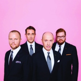 a quartet of men in fancy suits pose in front of a purple-pink background