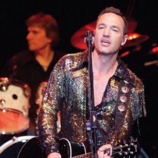 Man with dark short hair in a sparkly outfit with a guitar and a microphone singing. Member of the band is playing the guitar behind him.