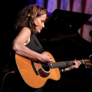 Woman with brown hair facing the right with a black sleeveless top on strumming a guitar. 