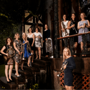 Nine women standing on a deck with stairs and railing. One woman standing in front of deck. All women are wearing dresses and smiling. Nine women of the ten women are holding instruments. 