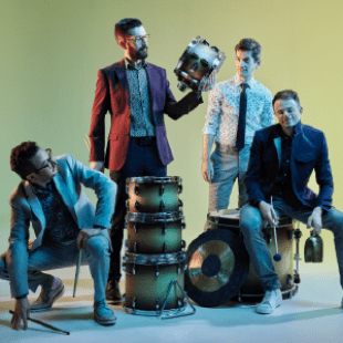 Four men in suits standing around a drum set that is not ensembled. One man is holding a cow bell. One man is holding drum sticks. One man is holding a piece of the drum set.