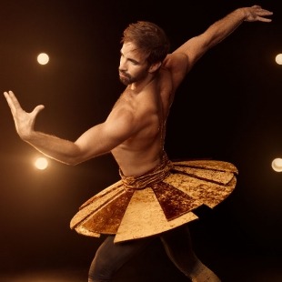 Man with dark hair and facial hair with no shirt and a yellow colored flat tutu. He is leaning to the side with his back arm  up and his front arm angled toward the ceiling.