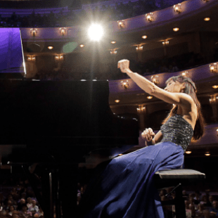 A woman sitting at a grand piano raises her right hand triumphantly with a large crowd watching her.  
