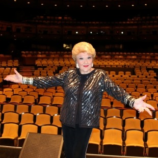 Woman in sparkly black jacket standing in front of theater seats with arms extended as if to welcome you to the theater