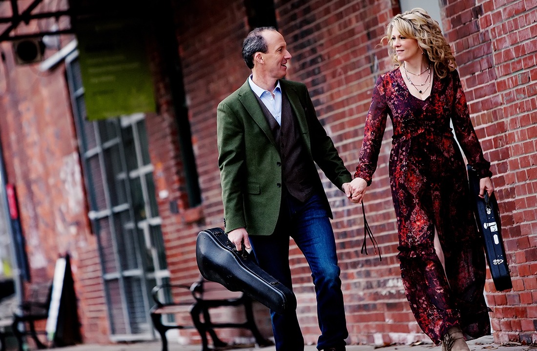Image of the two performers from Celtic Celebration holding hands and their fiddles in cases while walking by a brick wall.